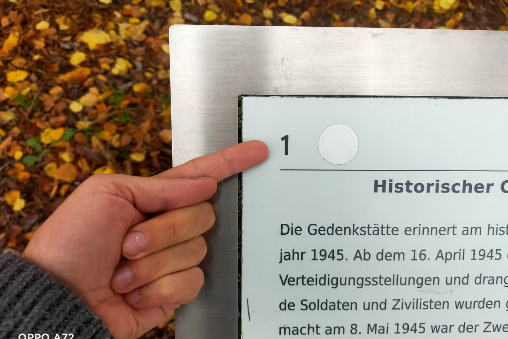 Ein Zeigefinger weist auf eine Nummer auf einer Info-Tafel im Außengelände der Gedenkstätte